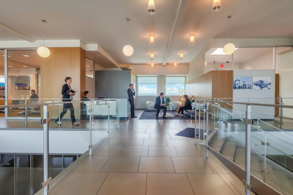 Barrister Suites Office Lease Modern office hallway with glass railings and tile floors. Several people in business attire are standing, walking, or sitting in small groups, engaging in conversation near large windows and contemporary light fixtures.