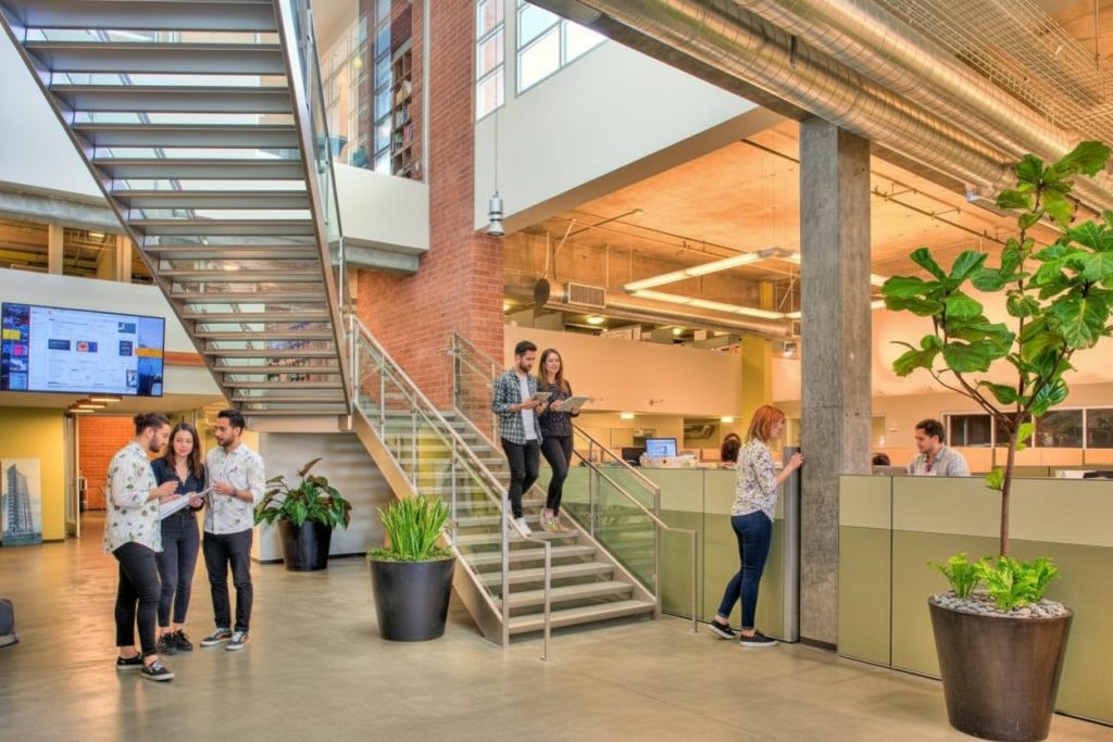 Barrister Suites Office Lease Modern open office with exposed ceilings and brick walls. Several people talk in groups near a glass staircase; others work at desks or write on a wall. Large potted plants decorate the bright, spacious workspace.