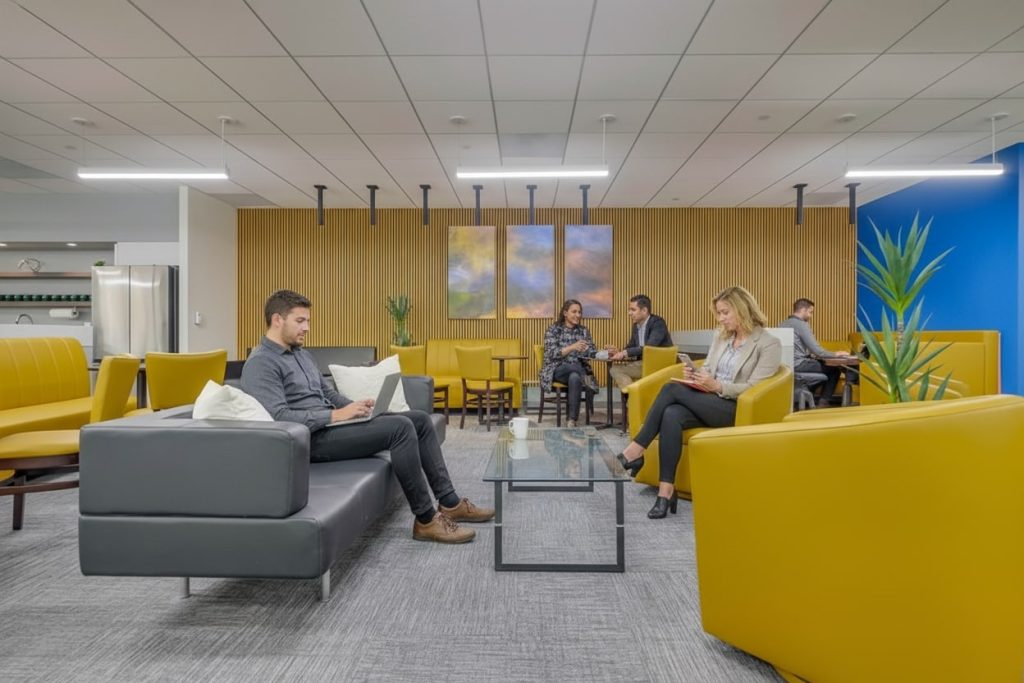 Barrister Suites Office Lease Modern office lounge with yellow and gray seating, glass coffee table, and several people working on laptops or having discussions. The background features wooden paneling and abstract wall art.