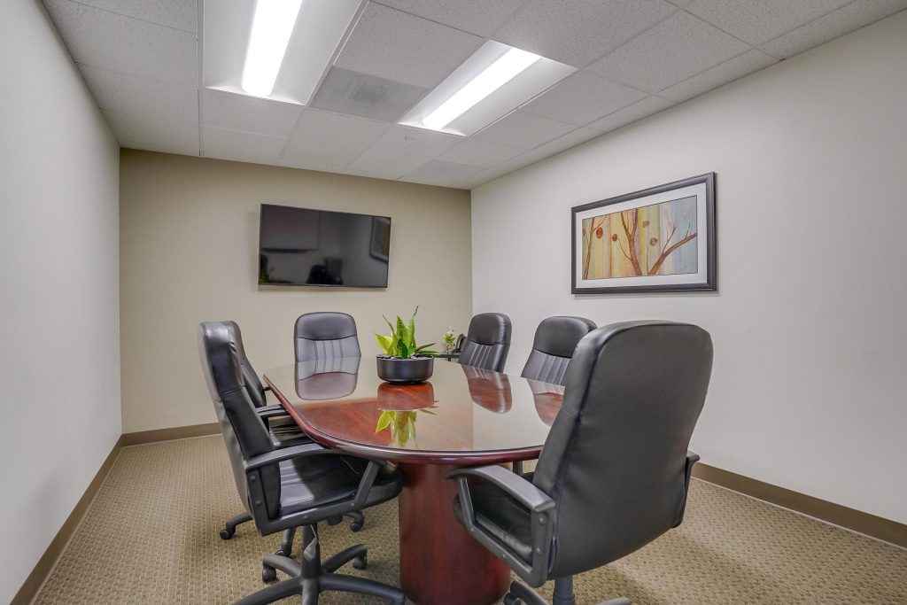 Barrister Suites Office Lease A small conference room with a polished oval table, six black office chairs, a wall-mounted TV, a framed abstract art piece, and a plant centerpiece on the table. The room has beige walls and carpet.