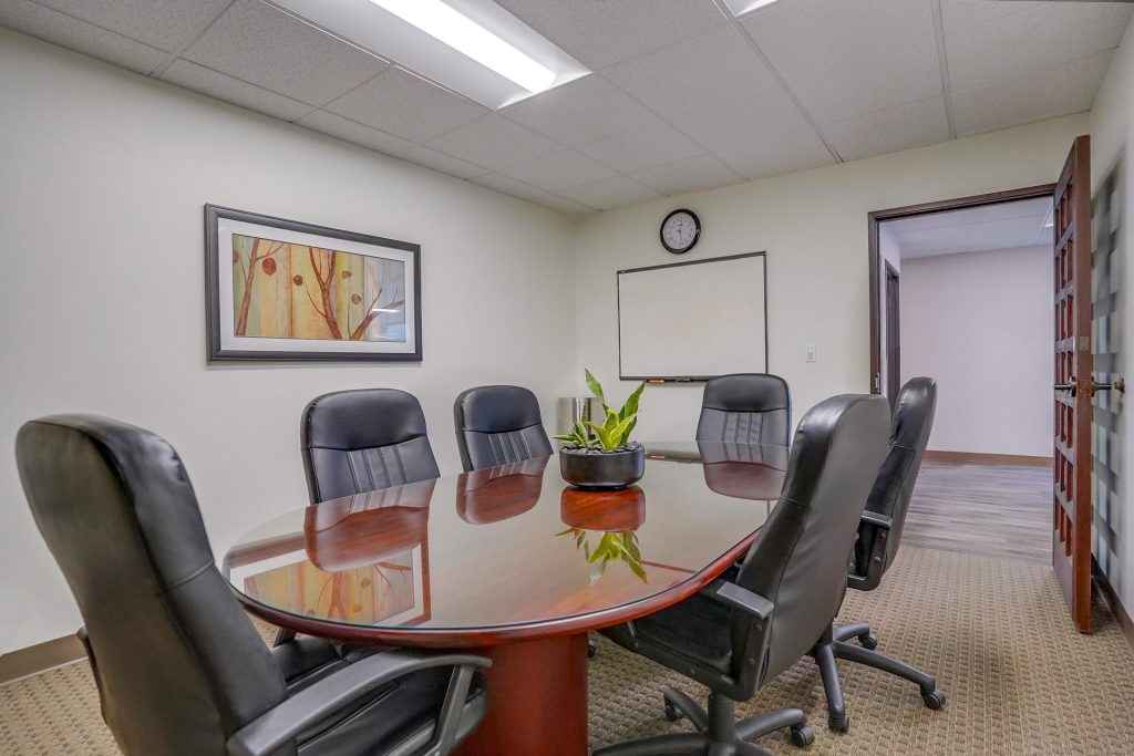 Barrister Suites Office Lease A small conference room with six black chairs around a polished wooden table, a potted plant in the center, a whiteboard, a clock, and framed artwork on the wall. The door is open to a hallway.