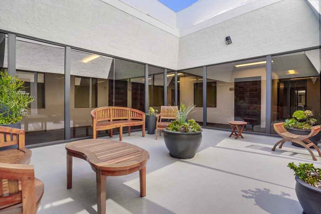 Barrister Suites Office Lease A modern outdoor courtyard with wooden benches and chairs, a few round tables, large potted plants, and glass walls reflecting the surrounding building under a clear blue sky.