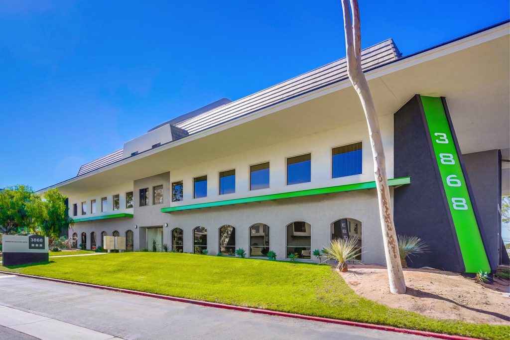 Barrister Suites Office Lease Modern two-story office building with large windows, green accents, and address number 3868 displayed vertically. The building is surrounded by a well-kept lawn and a few trees under a clear blue sky.