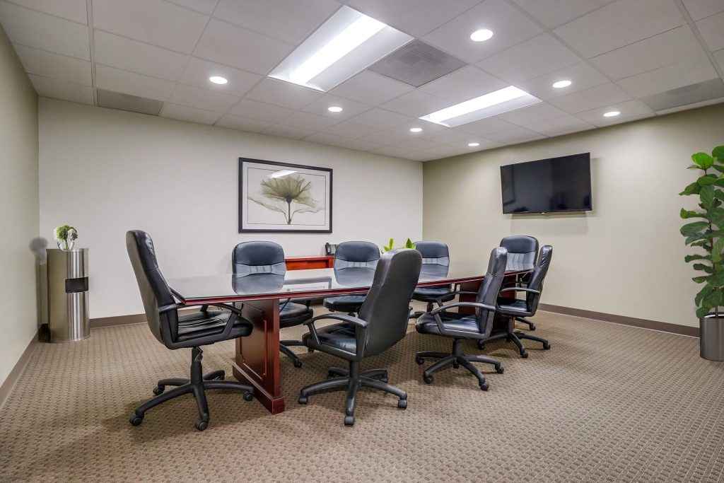 Barrister Suites Office Lease A modern conference room with a long wooden table, six black office chairs, a wall-mounted TV, a framed leaf artwork, potted plants, and neutral-colored walls and carpet.