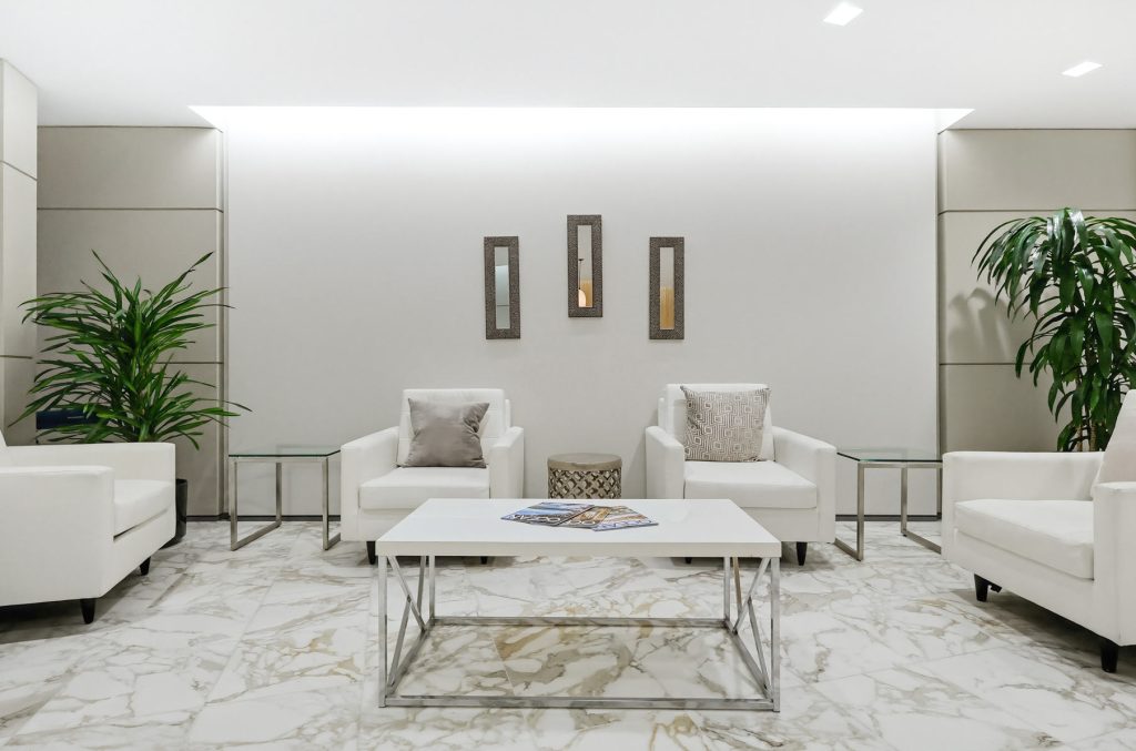 Barrister Suites Office Lease A modern waiting area with white armchairs, a marble coffee table, magazines, potted plants, and three rectangular mirrors on the wall, all set on a white marble floor.