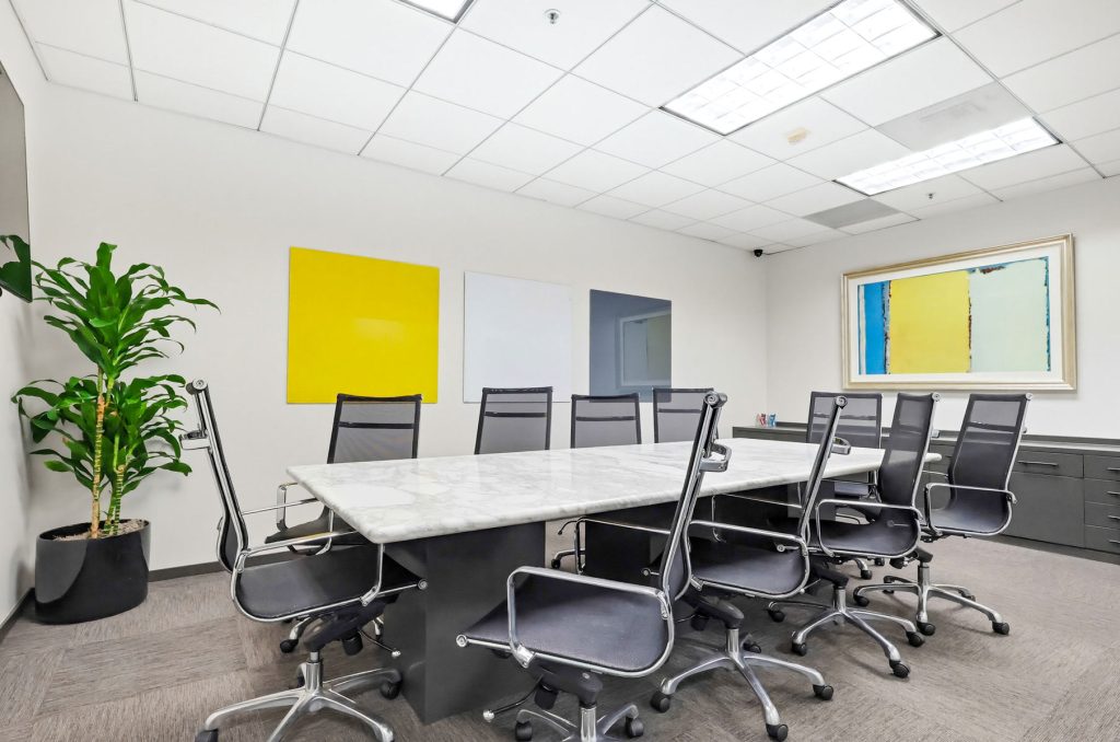Barrister Suites Office Lease A modern conference room with a long marble table, eight black office chairs, a potted plant, and colorful abstract art on white walls under bright ceiling lights.