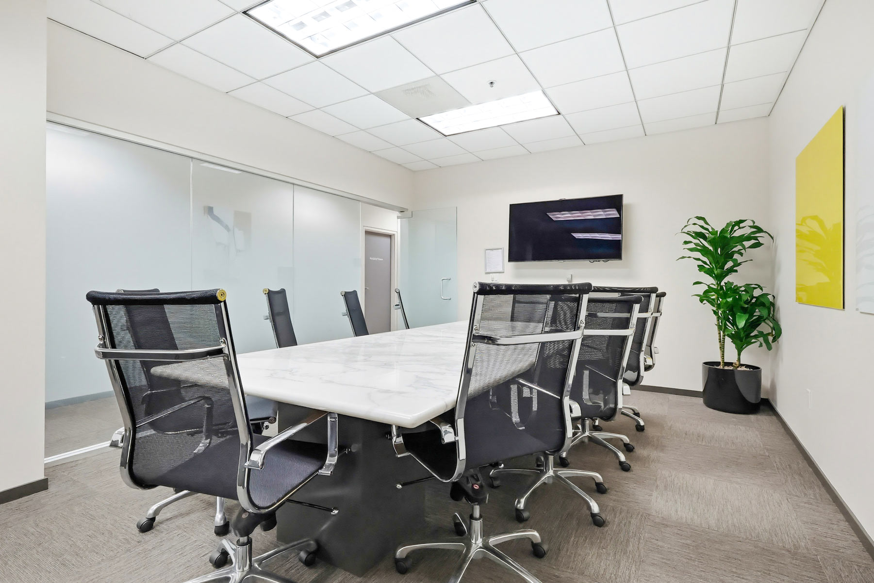 Barrister Suites Office Lease A modern conference room with a long marble table, eight black office chairs, a wall-mounted TV, a potted plant, white walls, abstract art, glass doors, and ceiling lights.