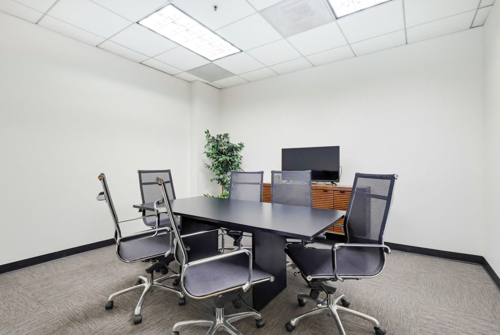 Barrister Suites Office Lease A small conference room with a black rectangular table, five mesh office chairs, a flat-screen monitor on a wooden cabinet, a potted plant in the corner, and white walls with a tiled ceiling.