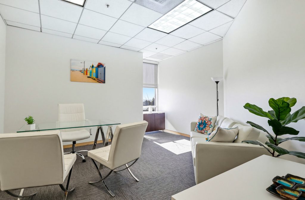 Barrister Suites Office Lease Modern office with white walls, glass desk, white chairs, a white sofa with a colorful pillow, a potted plant, and a beach painting on the wall. Large window lets in natural light.