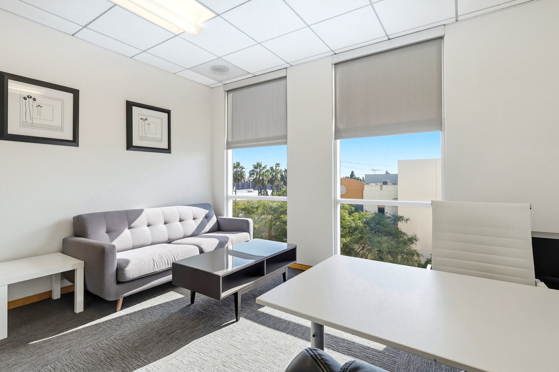Barrister Suites Office Lease A modern office with a gray sofa, white side table, black coffee table, white desk, white office chair, framed art on the wall, and large windows with shades letting in sunlight. Trees and buildings are visible outside.