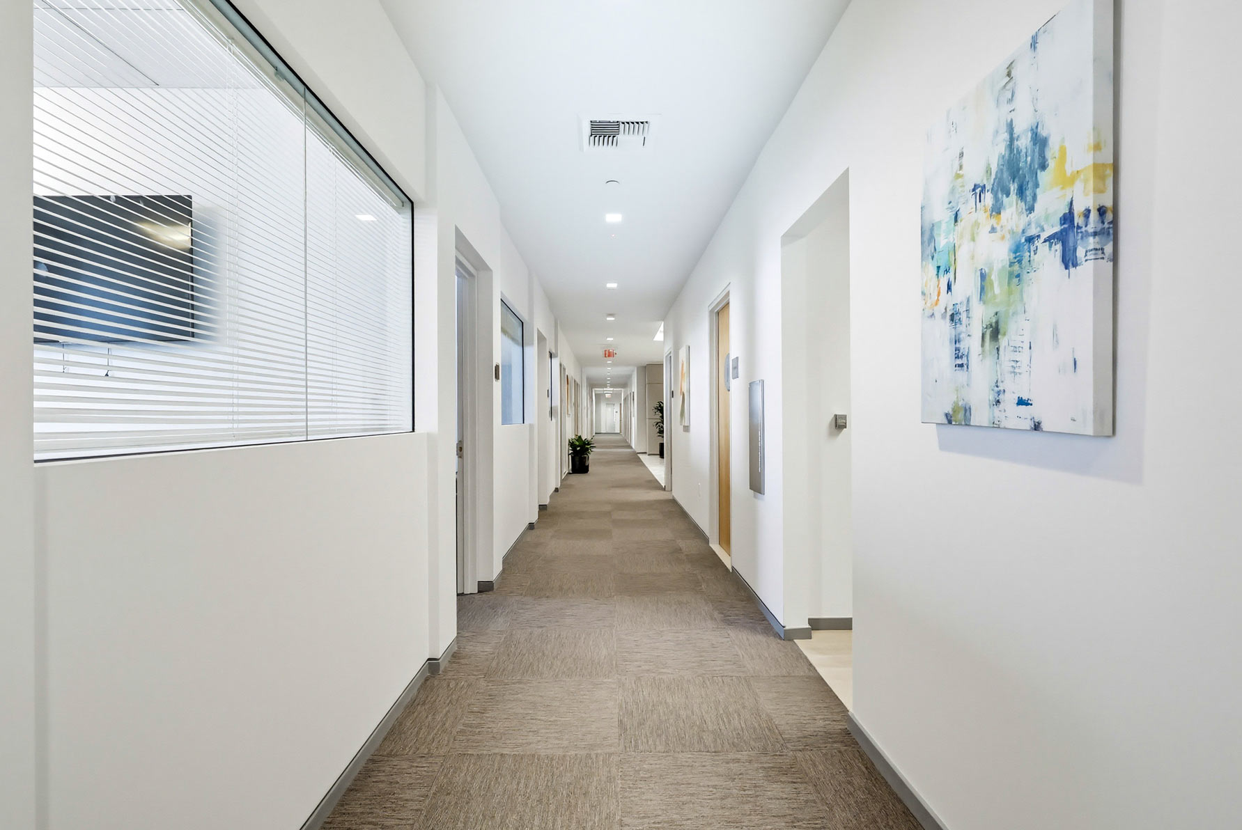 Barrister Suites Office Lease A clean, well-lit office hallway with beige carpet, white walls, several closed doors, modern abstract art on the wall, and a glass window overlooking another office space.