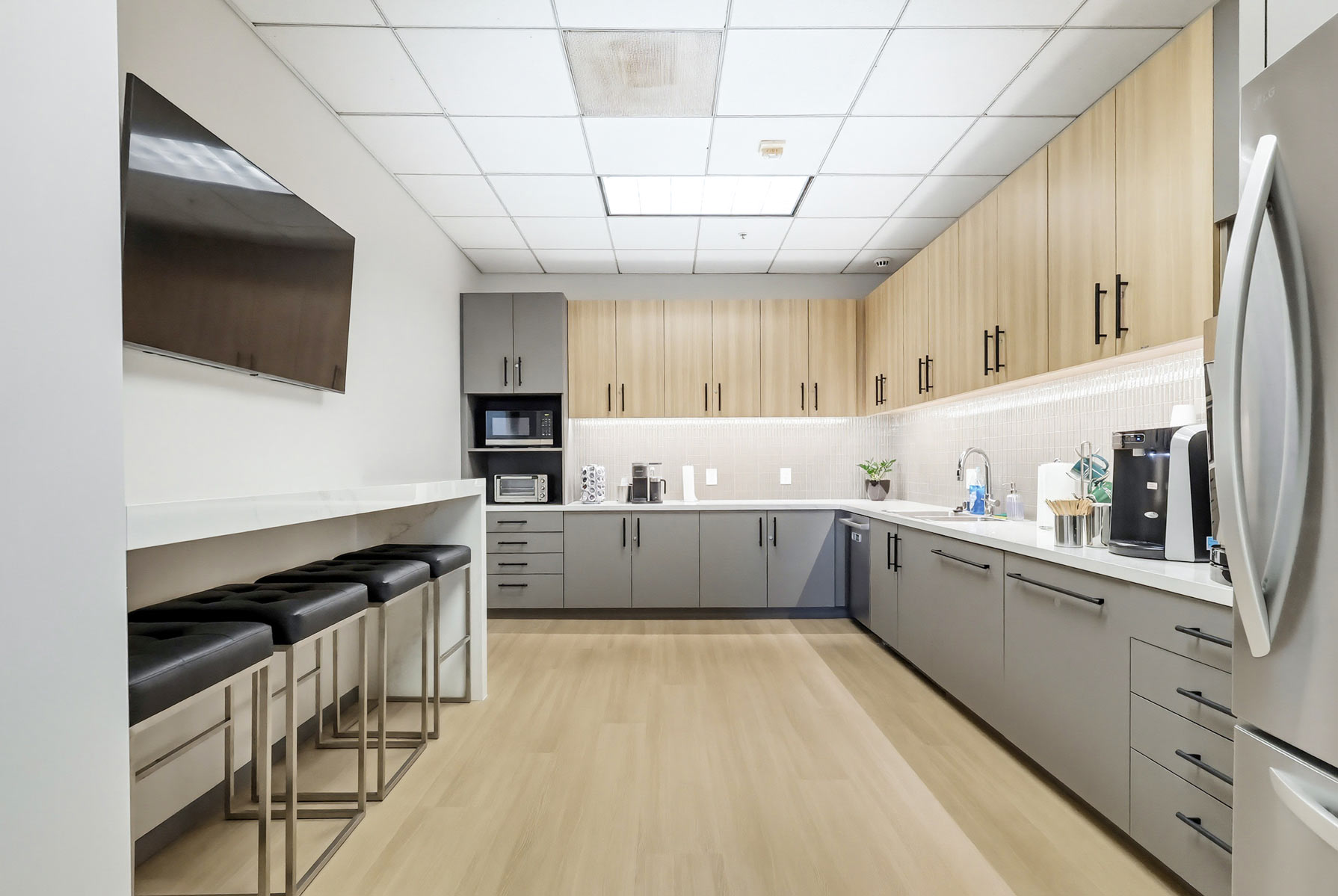 Barrister Suites Office Lease Modern office kitchen with light wood flooring, gray lower cabinets, light wood upper cabinets, a stainless steel fridge, a coffee maker, a microwave, a wall-mounted TV, and four black bar stools under a white counter.