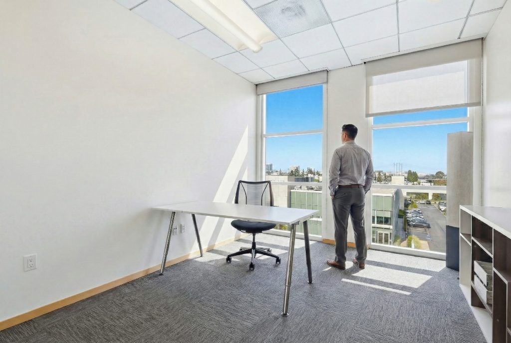 Barrister Suites Office Lease A man in business attire stands by large windows in a bright, modern office, looking outside. The office has a desk, chair, shelving unit, and neutral decor. Sunlight fills the space, highlighting its clean, minimalist design.