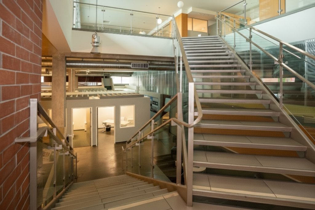 Barrister Suites Office Lease Modern indoor staircase with glass railings leads to an upper floor. The lower level shows open office spaces with lights on, white walls, and a brick wall on the left side. The area appears clean and well-lit.