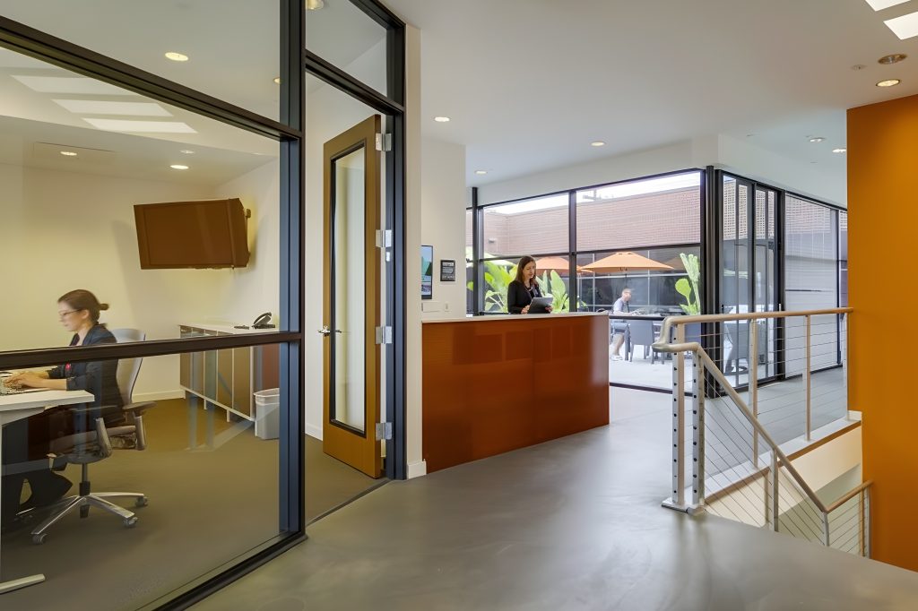 Barrister Suites Office Lease A modern office space with a glass-walled room on the left, a person working at a desk inside, and a reception area with another person at the counter. Large windows reveal an outdoor patio with tables and umbrellas.