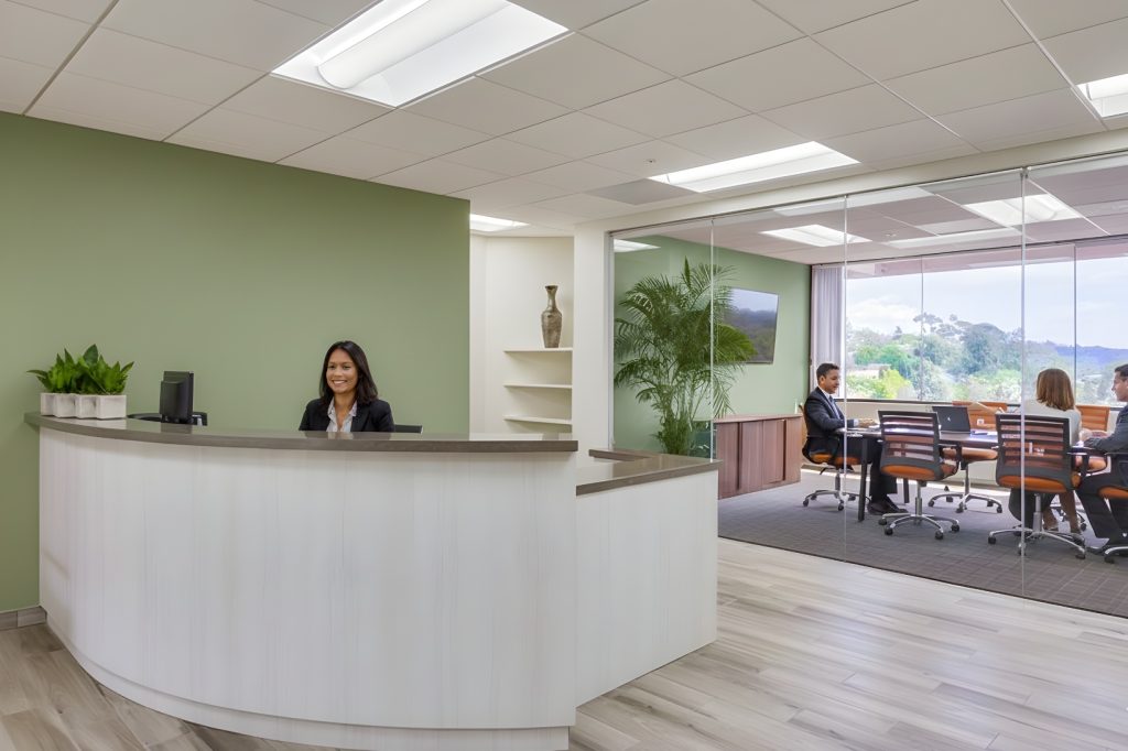 Barrister Suites Office Lease A modern office lobby with a smiling receptionist at a curved desk, green accent wall, and potted plants. Behind glass walls, several people are seated at a conference table in a meeting, with large windows showing a scenic outdoor view.