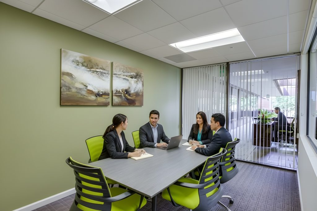 Barrister Suites Office Lease Four people in business attire sit around a modern conference table in a small office meeting room, engaged in discussion. A painting hangs on the wall, and large windows provide natural light.