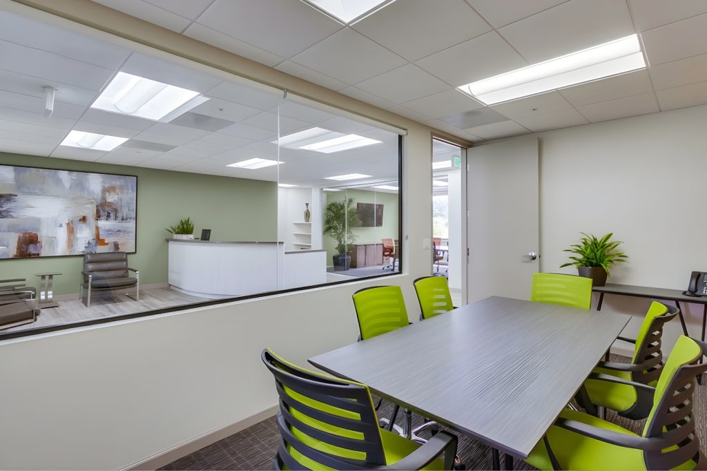 Barrister Suites Office Lease A modern office meeting room with a gray table, six green and black chairs, a potted plant, and large interior window overlooking a reception area with seating and artwork on the wall.