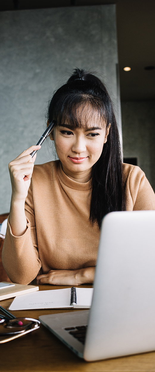 Barrister Suites Office Lease A woman with long dark hair in a ponytail sits at a table, holding a pen to her head, looking thoughtfully at a laptop. Papers, a notebook, and glasses are on the table in front of her.
