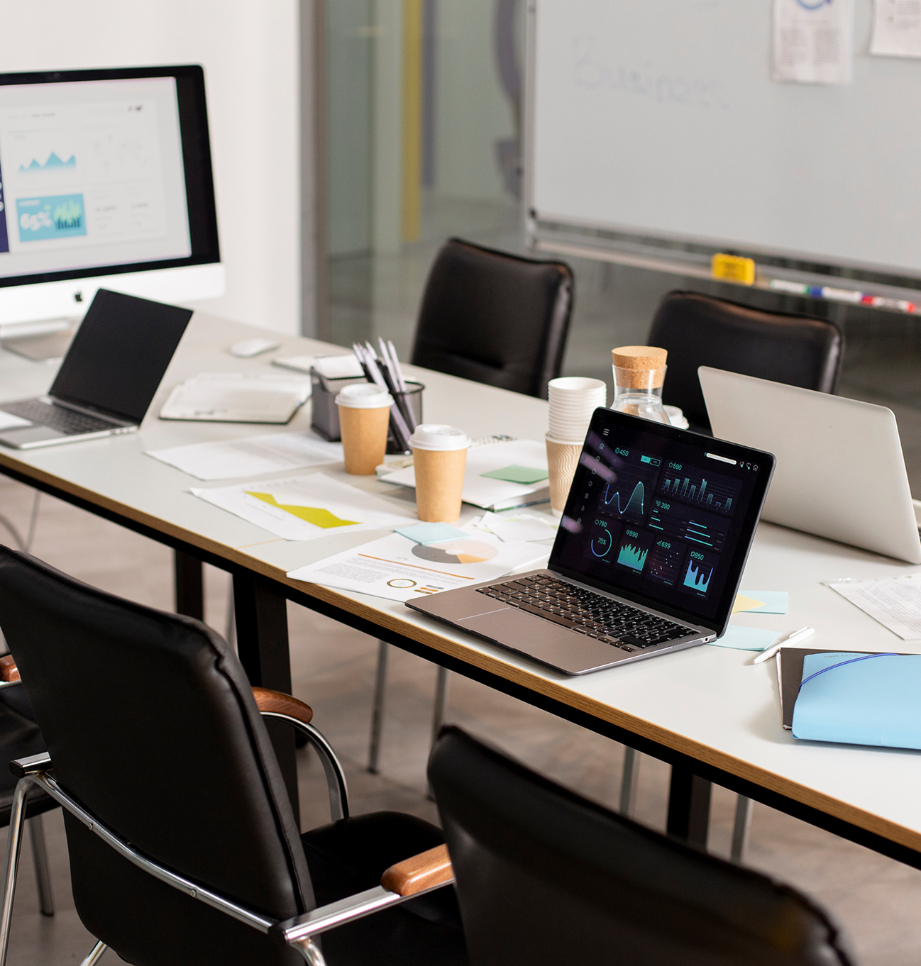 Barrister Suites Office Lease A modern conference room with a long table covered in laptops, documents, coffee cups, and papers. Several black chairs surround the table. A whiteboard and computer monitor are visible in the background.