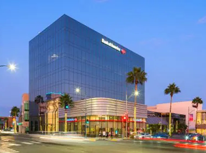Barrister Suites Office Lease A modern glass office building with a Bank of America sign, illuminated at dusk. Palm trees line the street, and light trails from passing cars are visible in the foreground.