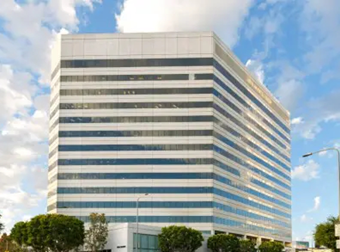 Barrister Suites Office Lease A modern, multi-story office building with reflective glass windows stands against a partly cloudy blue sky, flanked by a few trees and a streetlight in the foreground.