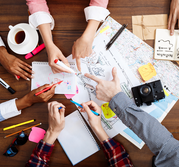 Barrister Suites Office Lease A group of people collaborate around a table with maps, notepads, pens, a camera, coffee, and sticky notes, planning a trip or project and pointing at destinations on the map.
