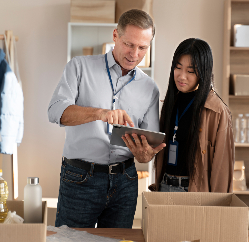 Barrister Suites Office Lease Two people, a man and a woman, stand together in a workspace, looking at a tablet. They wear name badges and are surrounded by cardboard boxes, suggesting a collaborative work or volunteer environment.