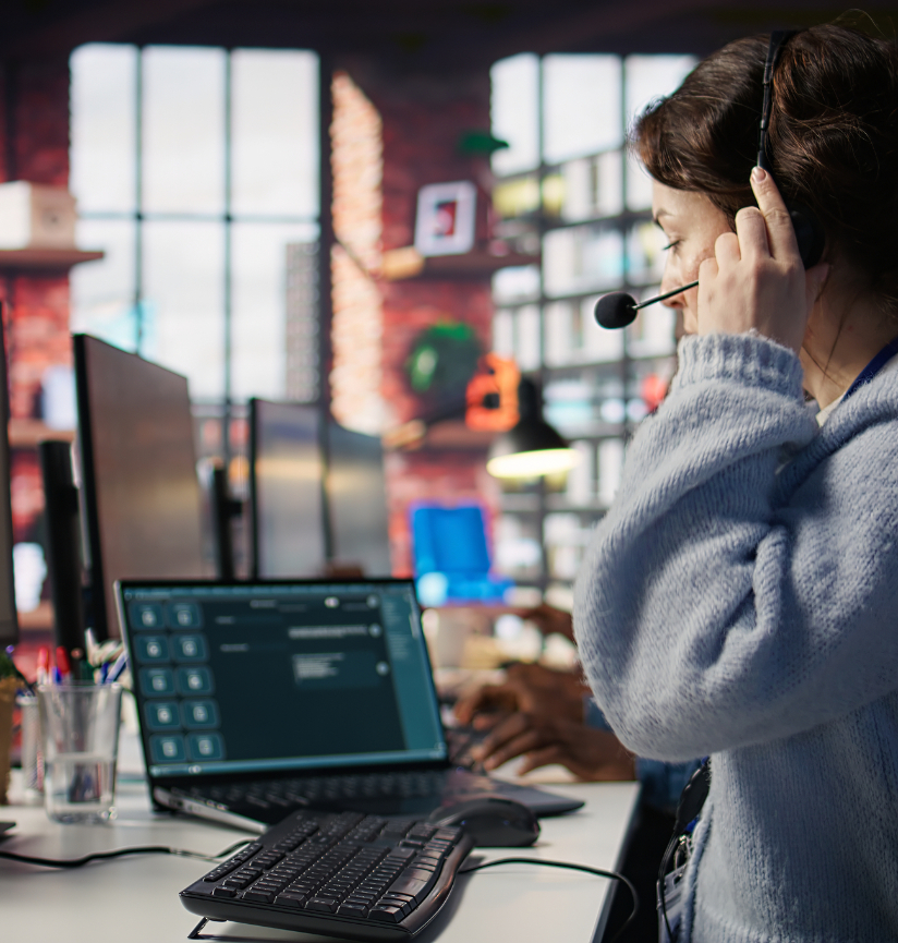 Barrister Suites Office Lease A woman wearing a headset sits at a desk with a laptop and keyboard, appearing to assist a customer. Computer screens and office supplies are visible, with a blurred office background and another person working nearby.