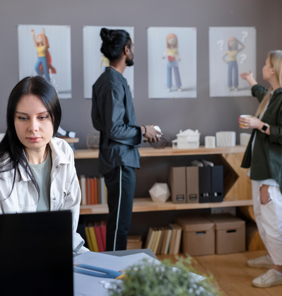 Barrister Suites Office Lease Four people in an office space; one woman works on a laptop in the foreground while three colleagues discuss and point at cartoon posters on the wall in the background. Shelves with files and decorative items are visible.