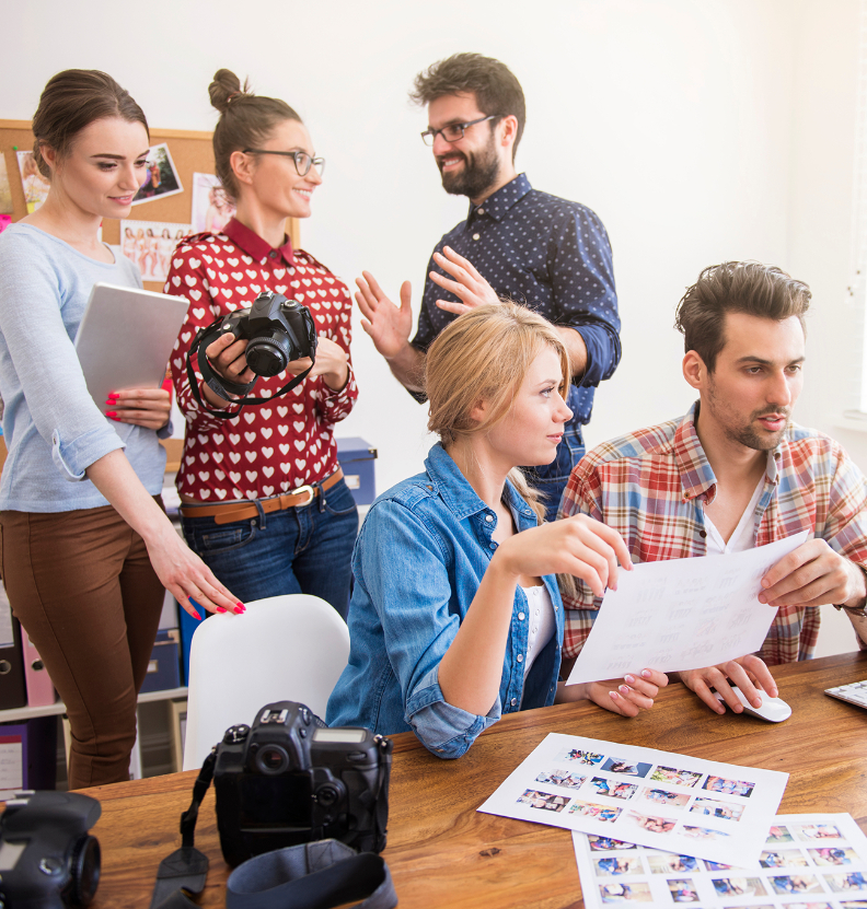 Barrister Suites Office Lease Five people collaborate in a bright office. Two are seated, looking at photos and a computer, while three stand nearby, holding a tablet and cameras, engaged in discussion. Cameras and photos are on the desk.
