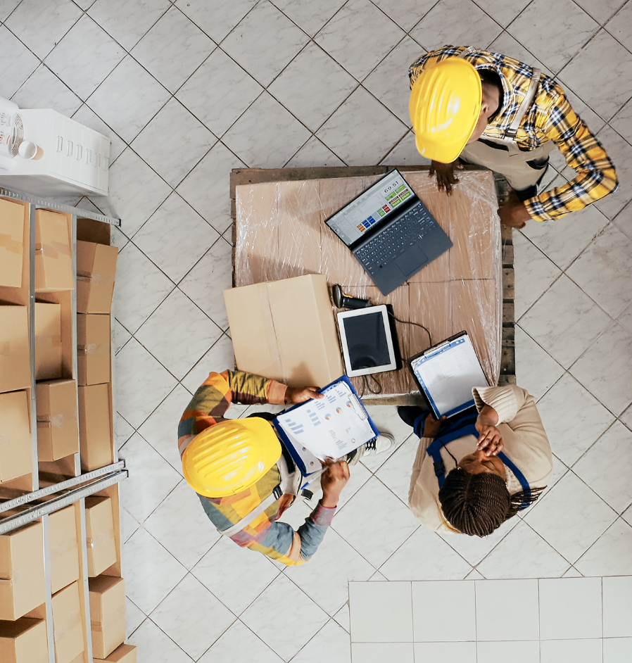 Barrister Suites Office Lease Three workers in yellow hard hats stand around a table with packages, a laptop, and clipboards, discussing inventory in a warehouse with shelves of cardboard boxes, viewed from above.