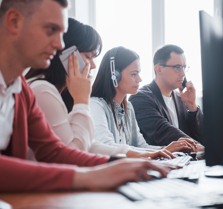 Barrister Suites Office Lease Four people sit in a row at desks using computers. Three are using phones, and one woman in the center wears a headset, all focused on their tasks in a bright office environment.