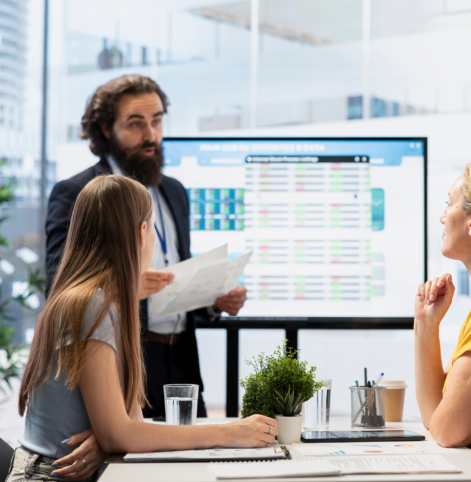 Barrister Suites Office Lease A man stands presenting data on a screen to two women seated at a conference table, with laptops, papers, and potted plants in a modern office setting.