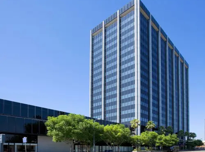 Barrister Suites Office Lease A tall modern office building with a glass exterior stands against a clear blue sky. Green trees line the street in front of the building, and a shorter building extends to the left.