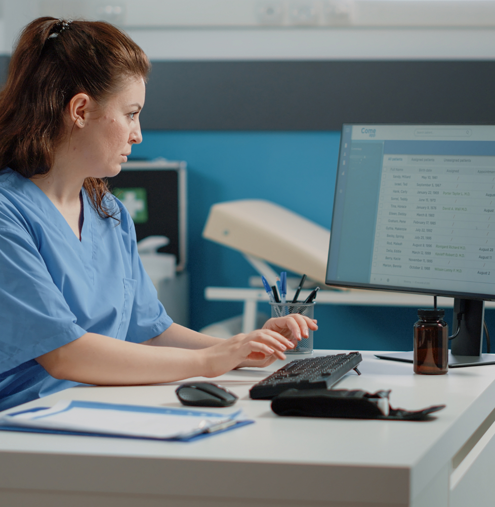 Barrister Suites Office Lease A healthcare professional in blue scrubs sits at a desk, typing on a keyboard and looking at a computer monitor displaying medical records. The desk has a clipboard, pen holder, brown bottle, and computer accessories.