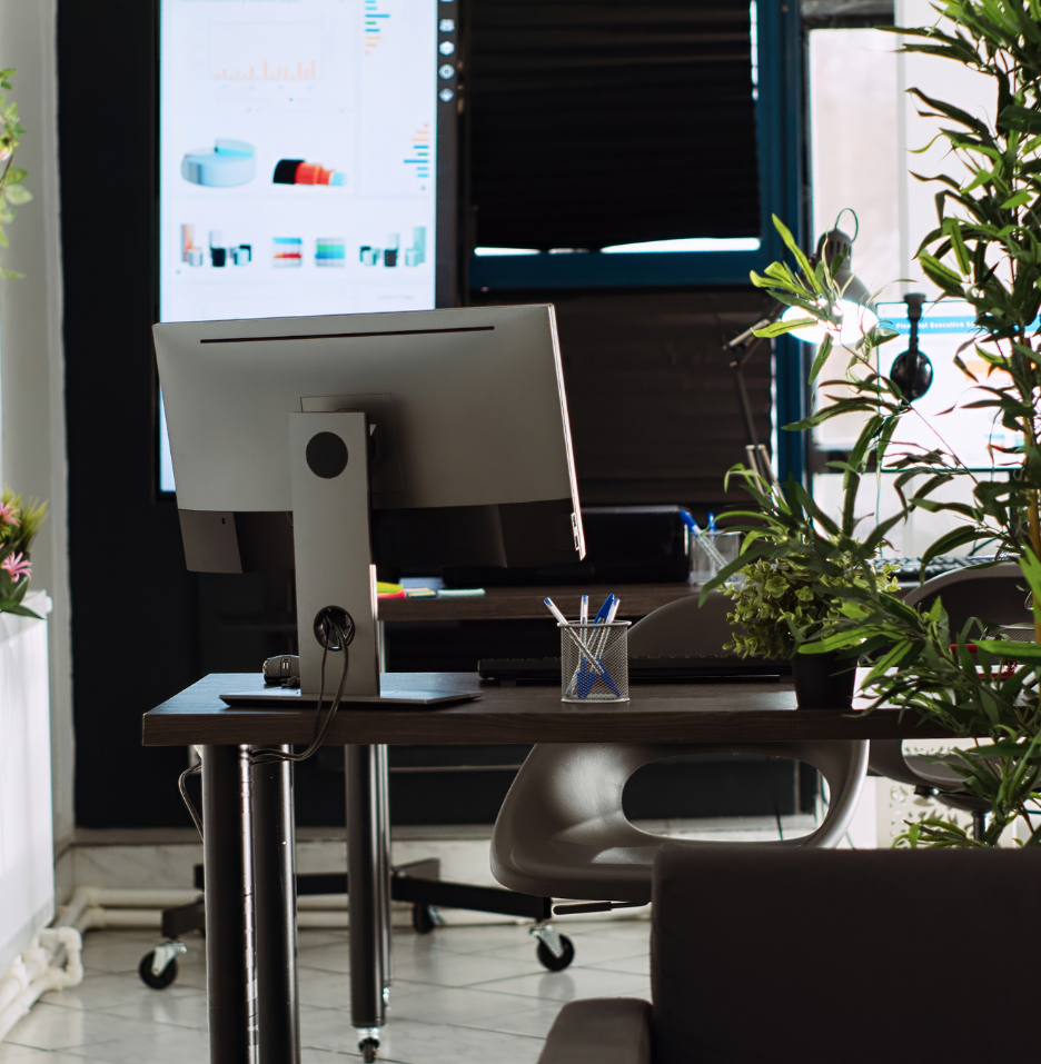 Barrister Suites Office Lease A modern office desk with a computer monitor, keyboard, mouse, and office supplies sits near large windows and green plants. A presentation with charts is displayed on a screen in the background.