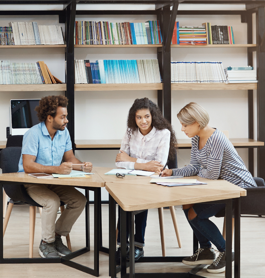 Barrister Suites Office Lease Three people sit at a table in a library or study area, writing and discussing work together. Shelves filled with books and magazines are visible in the background.
