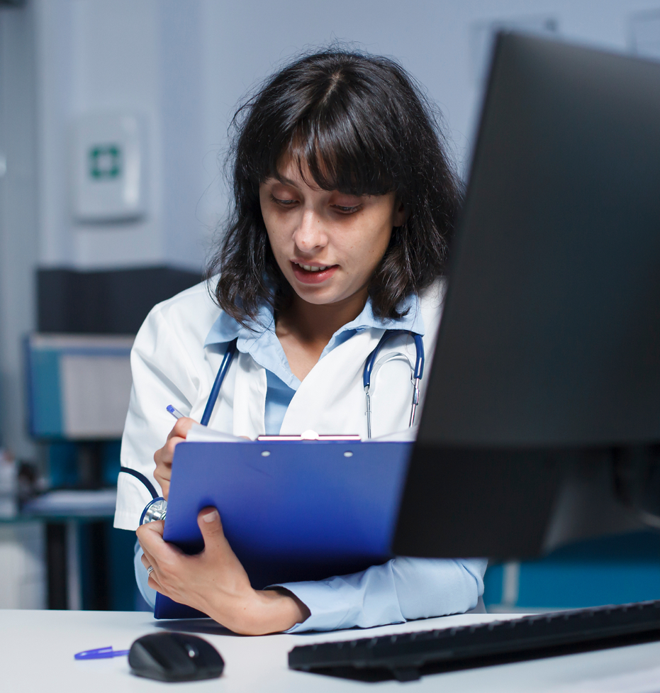 Barrister Suites Office Lease A female doctor with a stethoscope around her neck is sitting at a desk, writing on a blue clipboard while looking down, with a computer monitor and mouse in front of her.