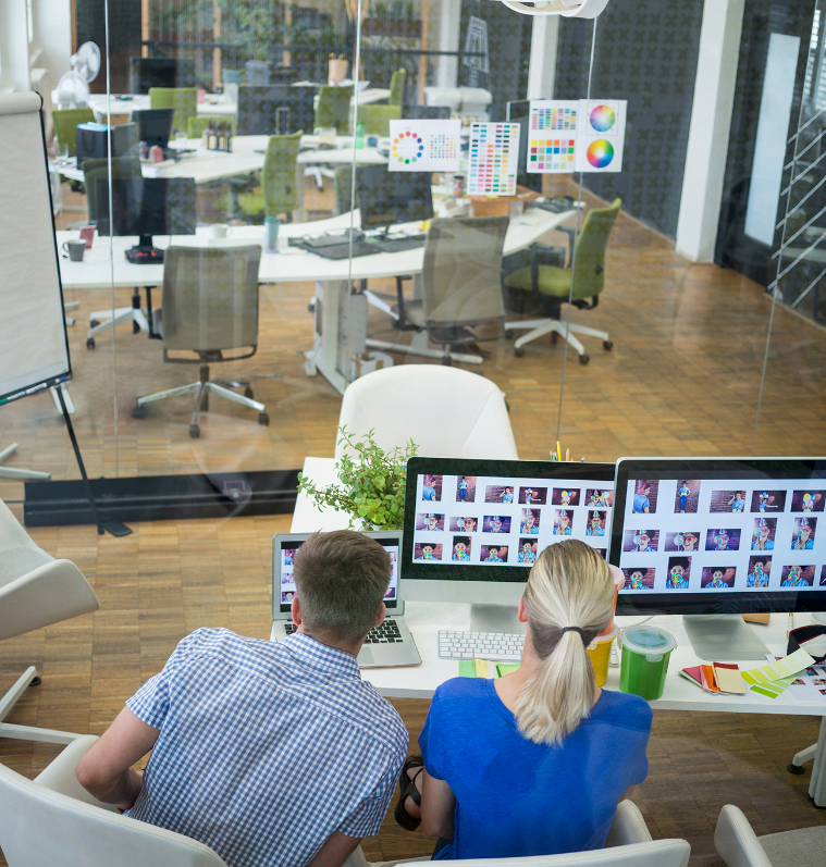 Barrister Suites Office Lease Two people sit at a desk, looking at computer monitors displaying image thumbnails in a modern office with glass walls and more empty desks and chairs in the background.