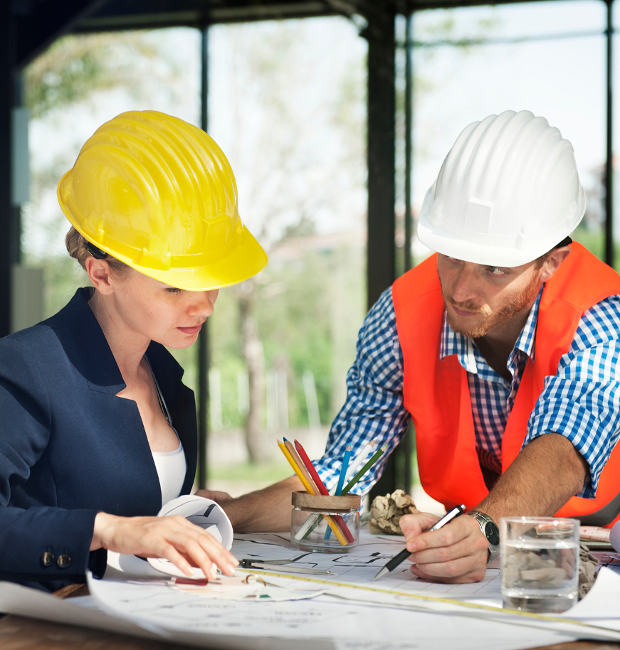 Barrister Suites Office Lease Two construction workers wearing hard hats review blueprints at a desk. The woman in a yellow helmet and blazer looks at plans, while the man in a white helmet and orange vest points and discusses details.