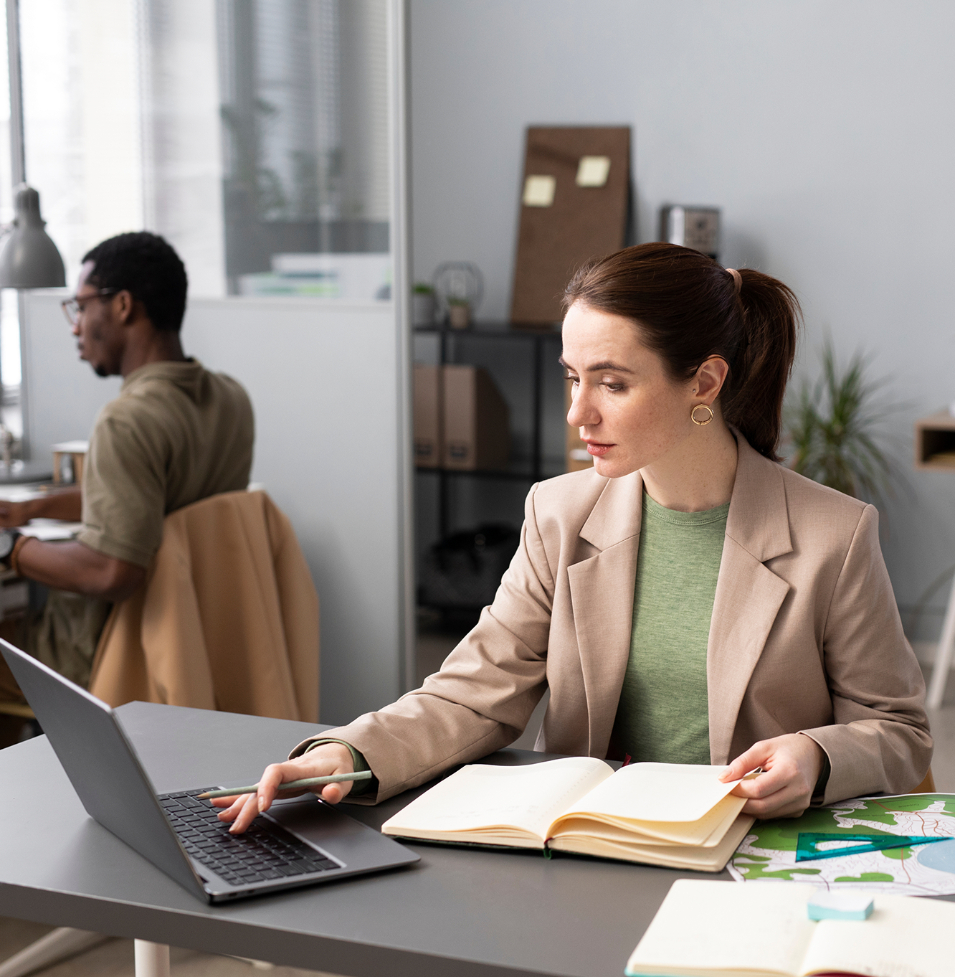 Barrister Suites Office Lease A woman in a beige blazer sits at a desk with an open notebook, using a laptop. She looks to the side thoughtfully. Papers and office supplies are on the desk. A man works at another desk in the background.