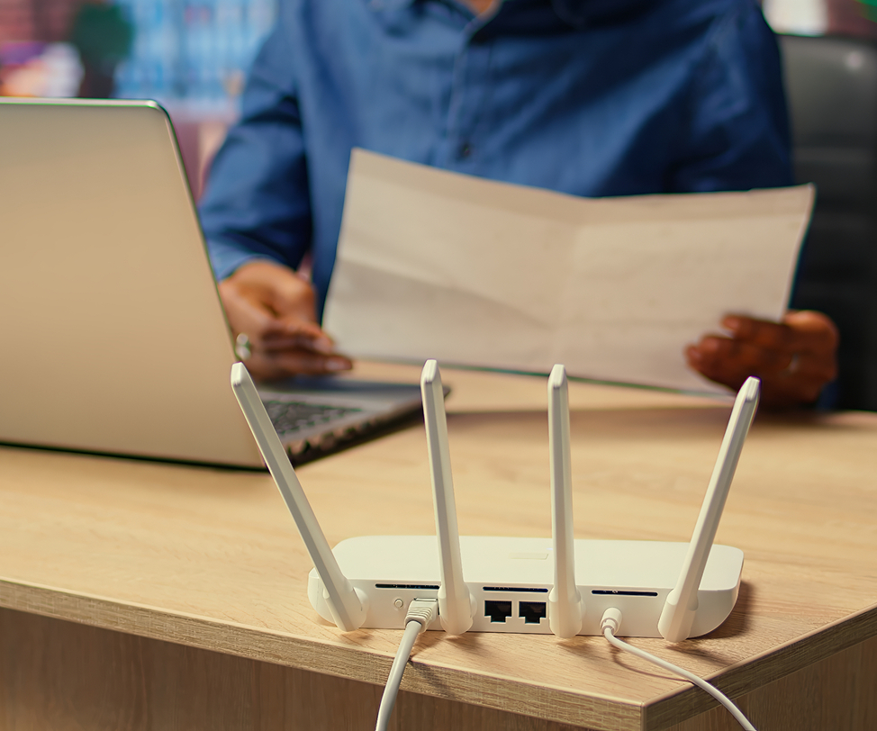 Barrister Suites Office Lease A person sits at a desk using a laptop and holding a sheet of paper. In the foreground, a white Wi-Fi router with four antennas and connected cables is in focus.