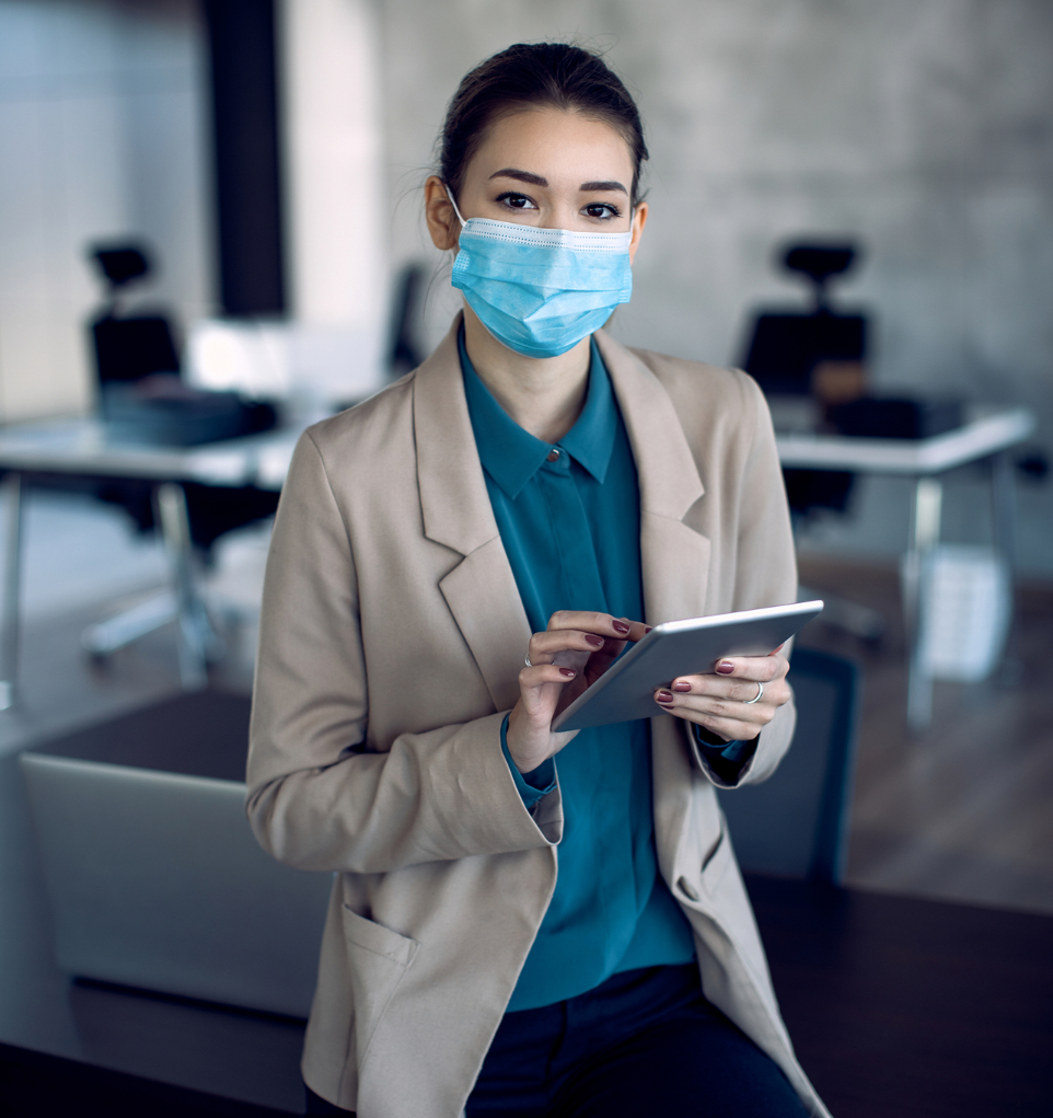 Barrister Suites Office Lease A woman wearing a face mask and a beige blazer holds a digital tablet in an office setting, with desks and chairs in the background.