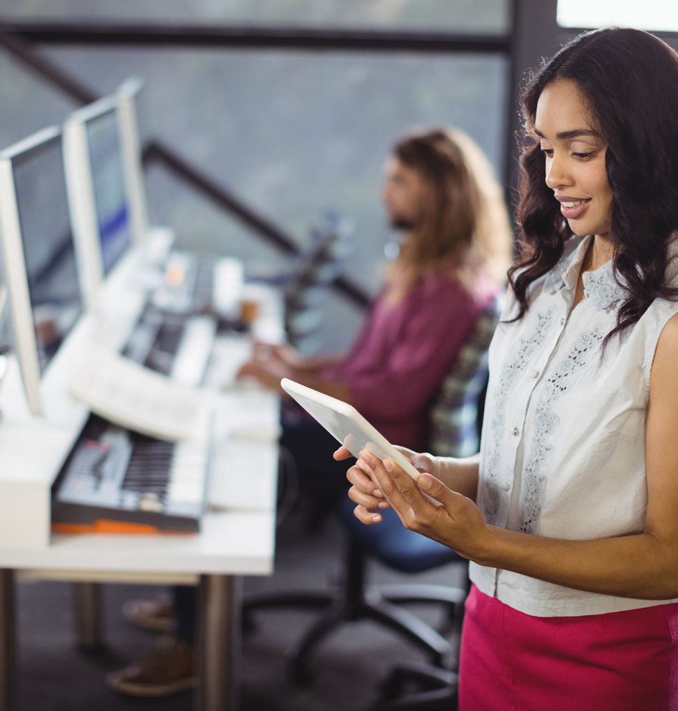 Barrister Suites Office Lease A woman stands in an office holding a tablet and smiling, while another person works at a computer with a keyboard and music equipment in the background.