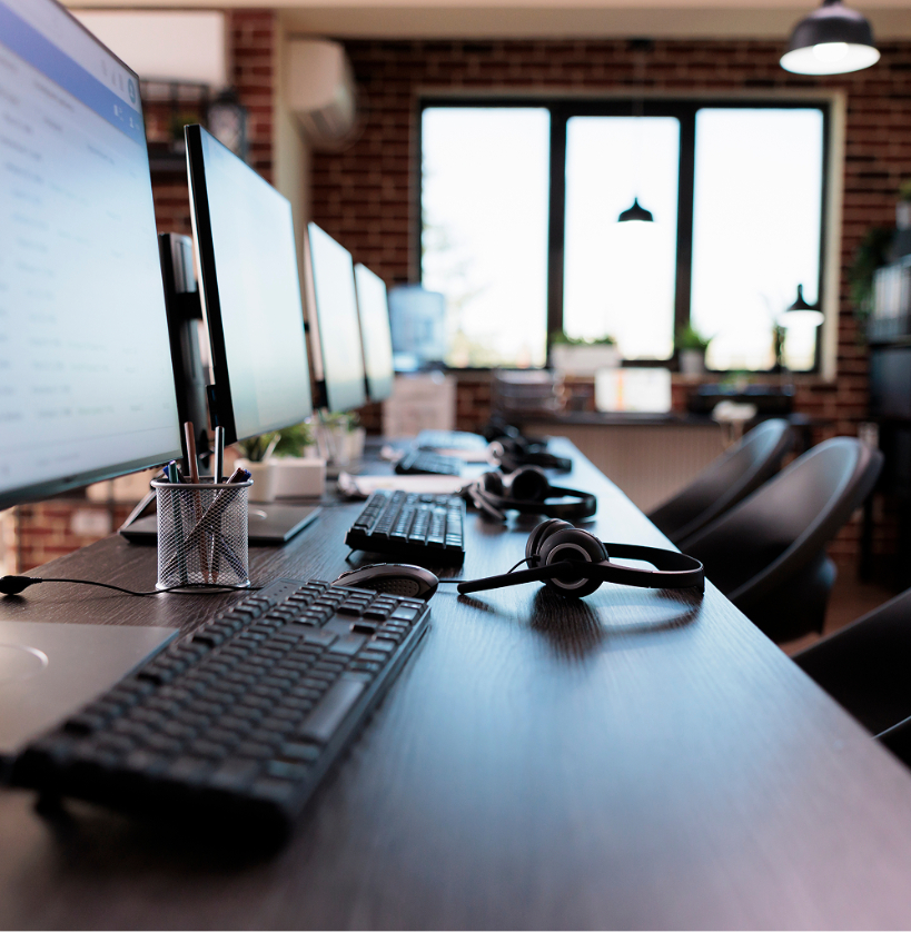 Barrister Suites Office Lease Row of computer monitors, keyboards, and headsets on a modern office desk, with chairs pushed in and large windows letting in natural light in the background.