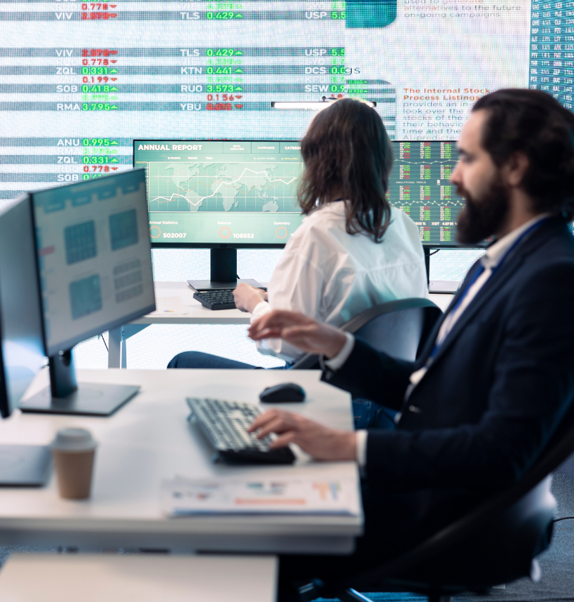 Barrister Suites Office Lease Two people work at computers in an office with large screens displaying stock market data and financial charts in the background. A coffee cup and documents are on a desk in the foreground.