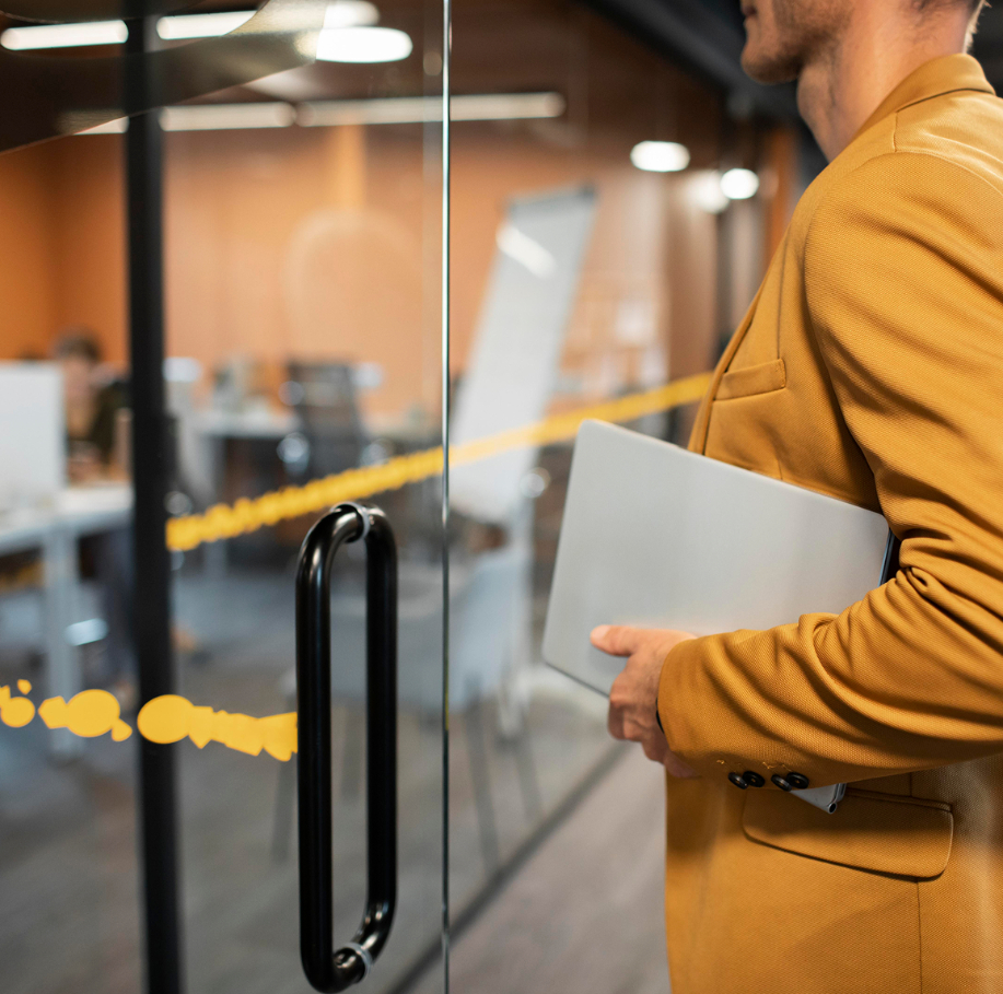 Barrister Suites Office Lease A person in a mustard-colored blazer holds a closed laptop while standing at the glass door of a modern office with desks and a whiteboard visible inside.