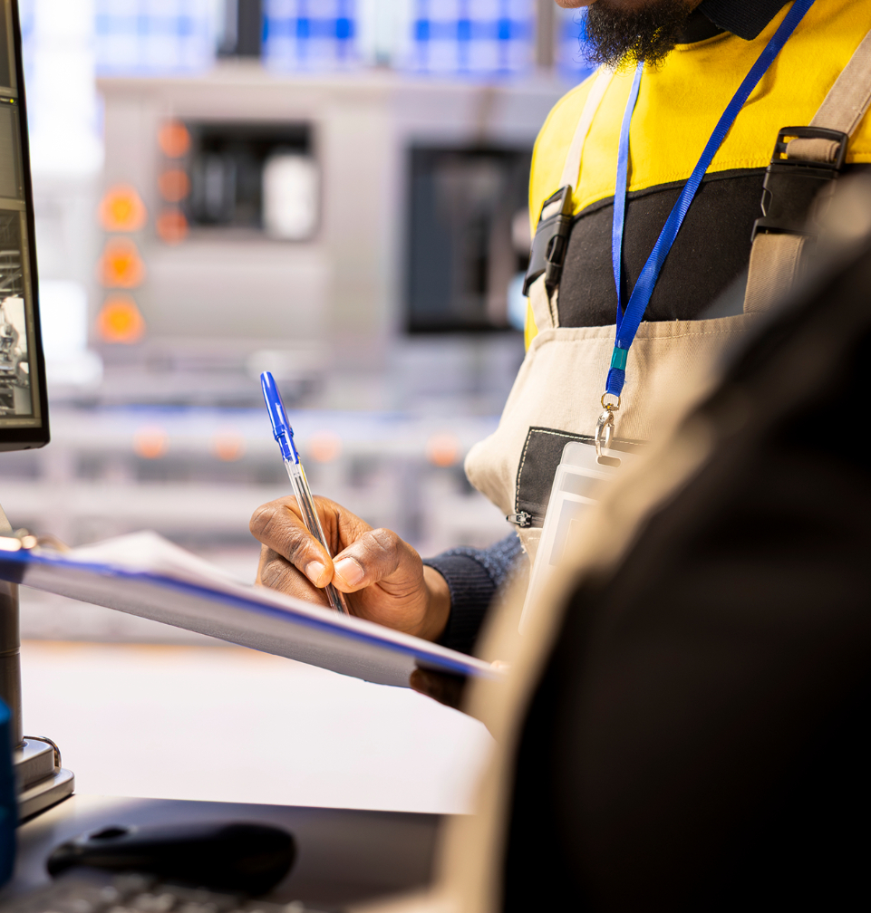 Barrister Suites Office Lease A person in work overalls and a yellow shirt writes on a clipboard with a blue pen, standing near a computer monitor in an industrial or factory setting. A lanyard with an ID badge is visible around their neck.
