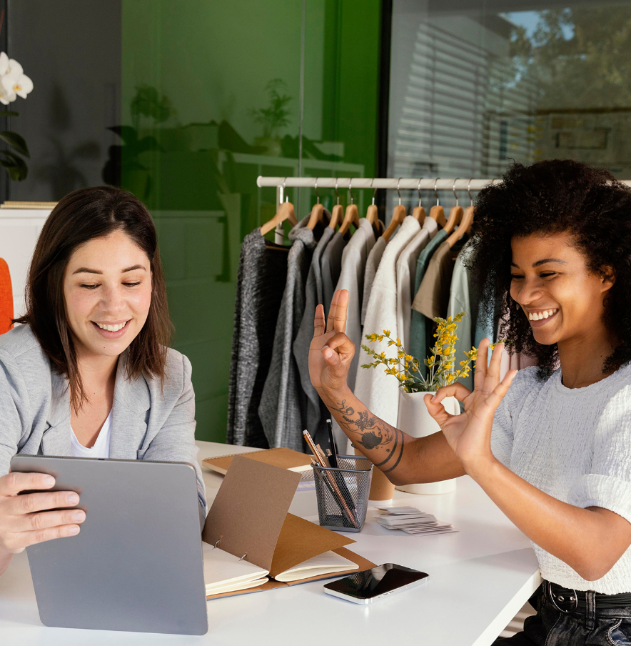 Barrister Suites Office Lease Two women sit at a desk in a modern office, smiling and looking at a tablet. One gives an “OK” hand gesture. Clothes hang on a rack behind them, suggesting a fashion or retail workspace.